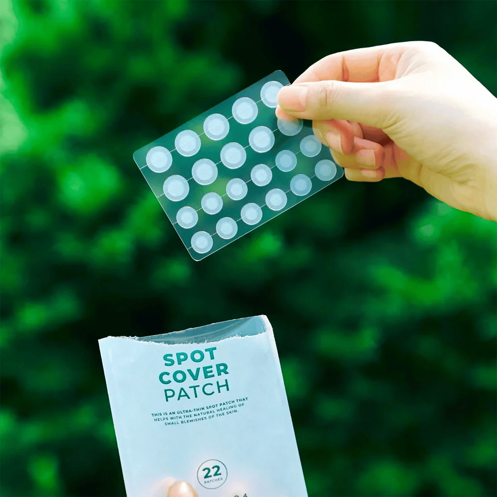 En hånd holder opp et ark med Spot Cover Patches, som er gjennomsiktige og avrundede. I bakgrunnen sees et uskarpt grønt miljø. Forpakningen viser en del av forpakningen med teksten "SPOT COVER PATCH" og informasjon om at produktet inneholder 22 plaster som hjelper til med å helbrede små hudområder.