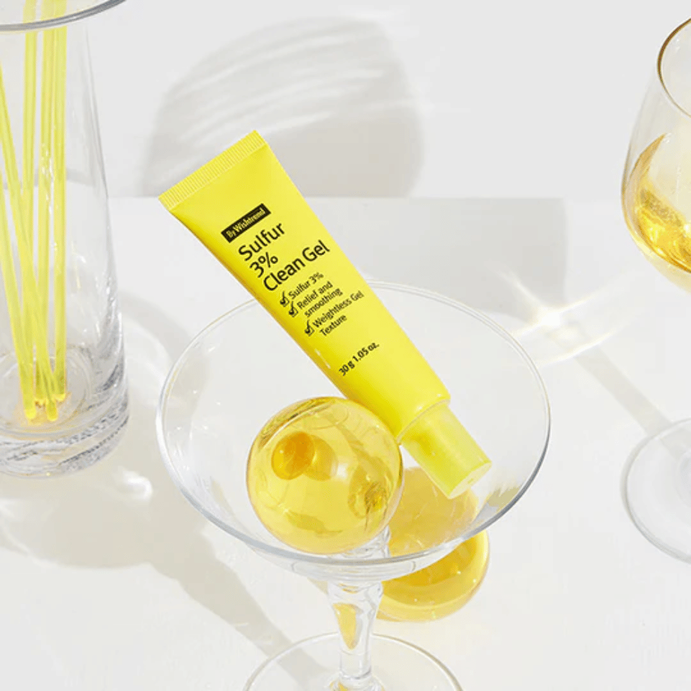 By Wishtrend Sulfur 3% Clean Gel i en gul tube plassert i et cocktailglass med en gul sfære, omgitt av elegante glass på en hvit bakgrunn.