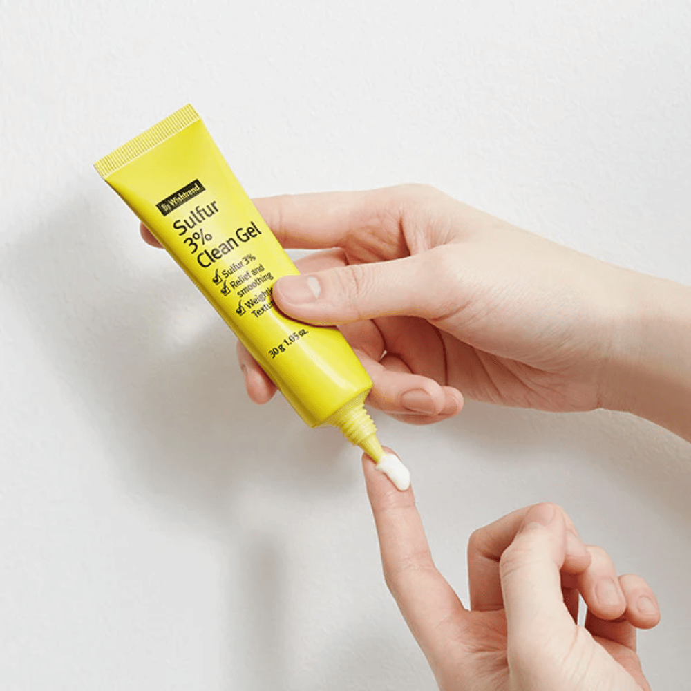 Nærbilde av hender som påfører By Wishtrend Sulfur 3% Clean Gel, en gul tube med hvit gel, mot en hvit bakgrunn.