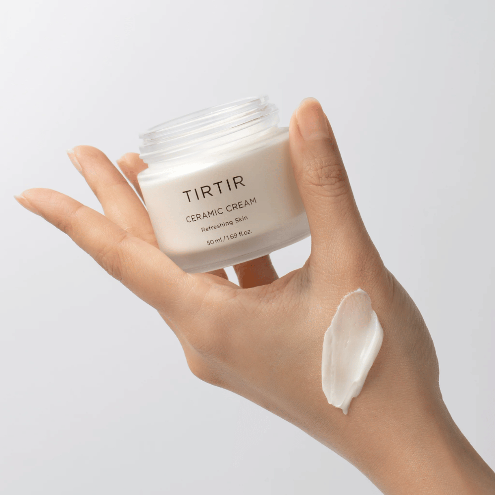 En hånd holder en boks med TIRTIR Ceramic Cream. På håndryggen er en mengde av kremen påført. Kremen ser ut til å være tykk og fuktighetsgivende. Emballasjen angir at innholdet er for "Refreshing Skin" og mengden er 50 ml / 1.69 fl oz.