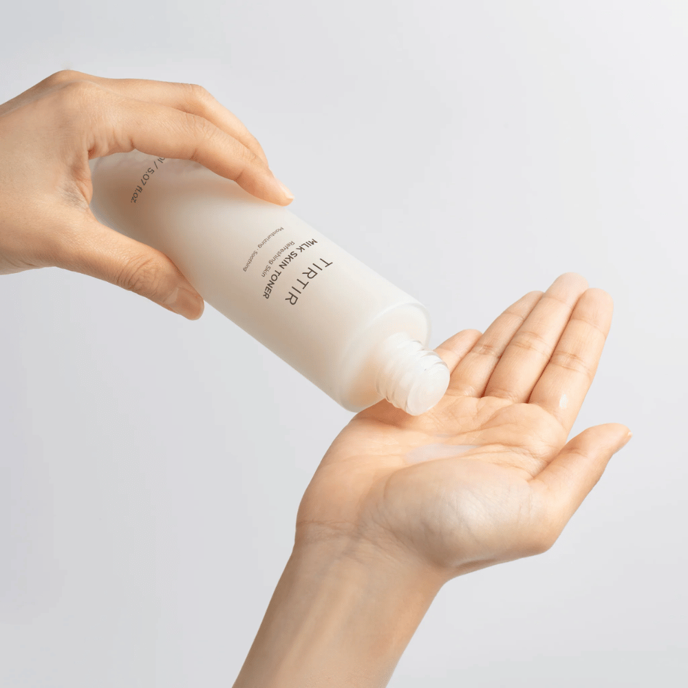 Bildet viser en person som heller ut en melkeaktig væske fra en flaske merket "TIRTIR Milk Skin" på håndflaten. Produktet ser ut til å være en hudtoner eller fuktighetsgivende lotion.