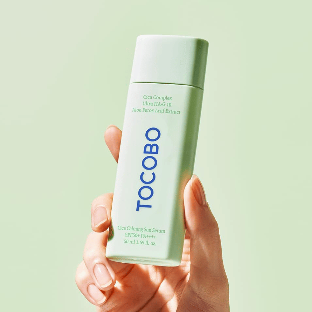 Hånd holder i TOCOBO Cica Calming Sun Serum med mintgrønn emballasje og blå tekst mot lysegrønn bakgrunn.