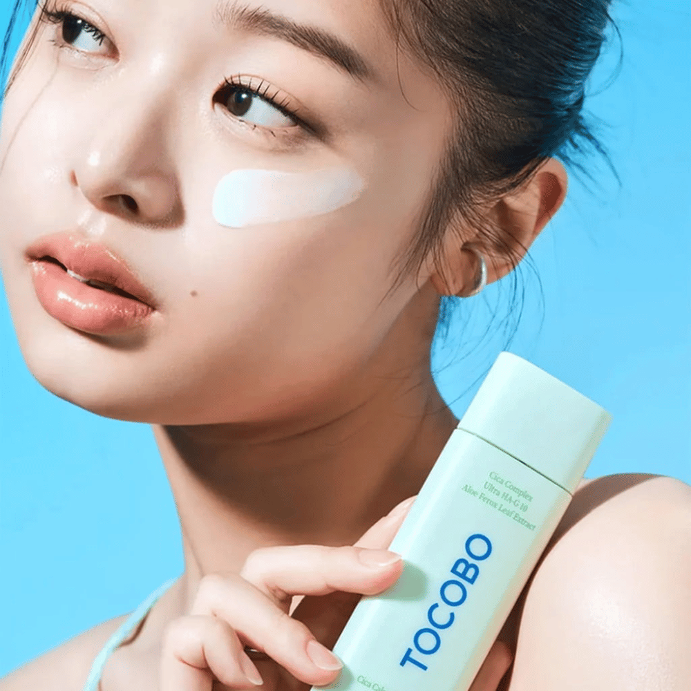 Modell med solkrem under øyet holder i TOCOBO Cica Calming Sun Serum i mintgrønn emballasje mot blå bakgrunn.