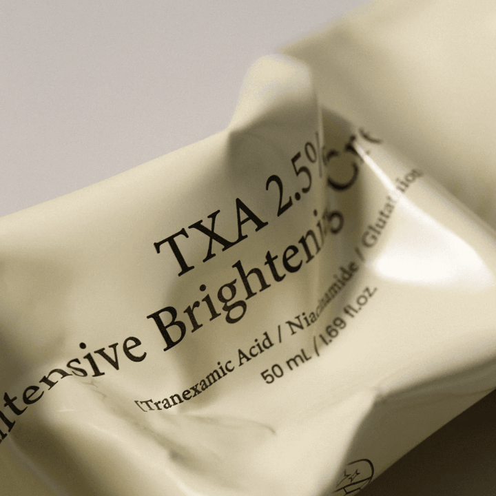 Nærbilde av AXIS - Y TXA 2.5% Intensive Brightening Cream der det myke og minimalistiske tubedesignet står i fokus. Bildet fremhever produktnavn, Tranexamic Acid og Niacinamide samt den elegante beige emballasjen.