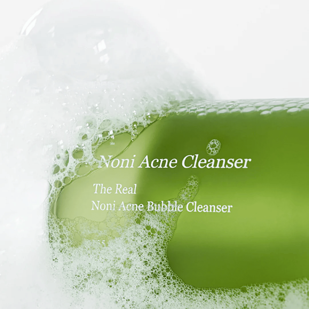 Flasken med The Real Noni Acne Bubble Cleanser er i fokus og omgitt av rikelig med skum. Den friske grønne fargen signaliserer naturlige ingredienser, perfekt for å rengjøre og bekjempe akne. Et ideelt produkt for en ren og klar hud.