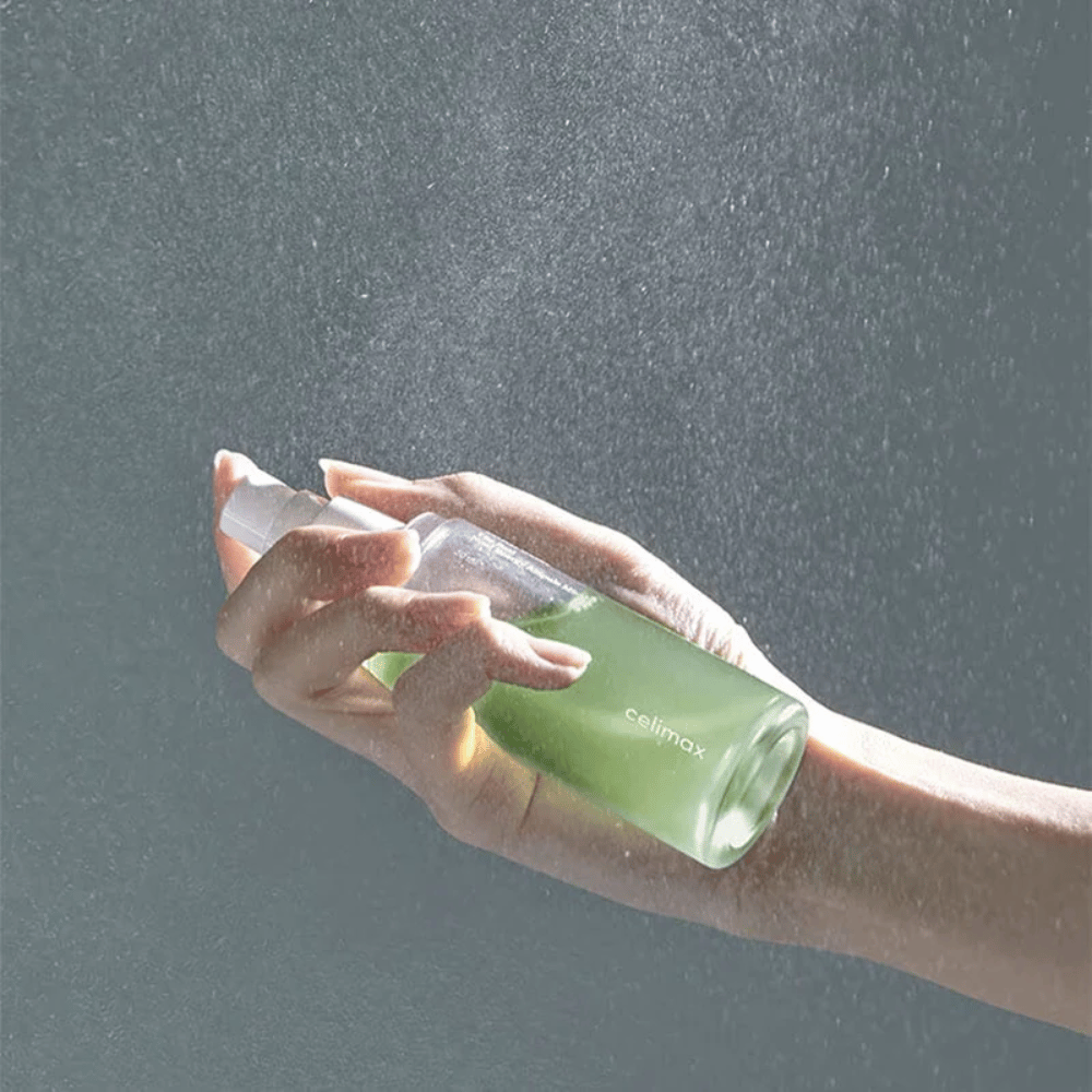 En hånd holder Celimax Noni Mist sprayflaske og påfører produktet i en fin, jevn spray på huden. Flasken er grønn og produktet markedsføres som "The Real Noni Energy Ampule Mist" for å gi energi og fuktighet til huden.