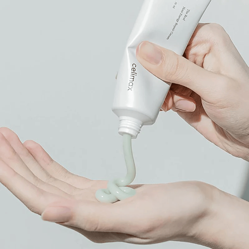 En hånd holder en hvit tube med Celimax Noni Energy Repair Cream, og kremen presses ut i en jevn stråle på håndflaten. Kremen er lysegrønn og markedsføres for å gi næring og gjenopprette huden.