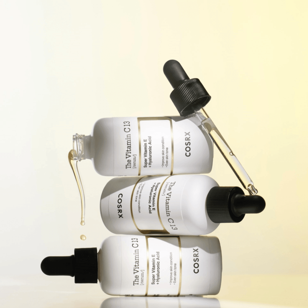 COSRX The Vitamin C 13 Serum med vitamin E og hyaluronsyre, tre flasker med pipette og dråper av C-vitaminserum mot ujevn hudtone og glød.