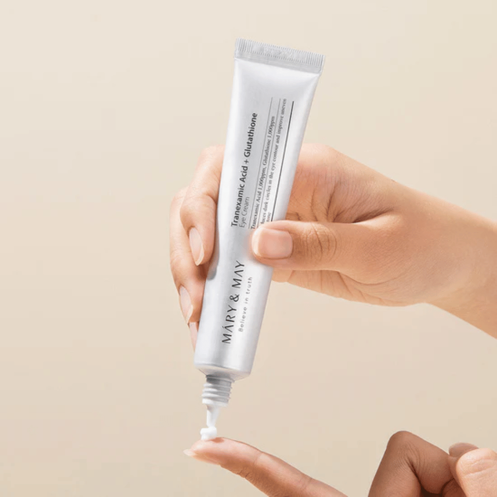 En hånd holder en tube MARY & MAY "Tranexamic Acid + Glutathione Eye Cream". Tuben er hvit og designen er ren og enkel, noe som fremhever produktets beroligende og lysnende egenskaper for øyeområdet. Produktet påføres forsiktig, med fokus på presisjon og effektivitet.