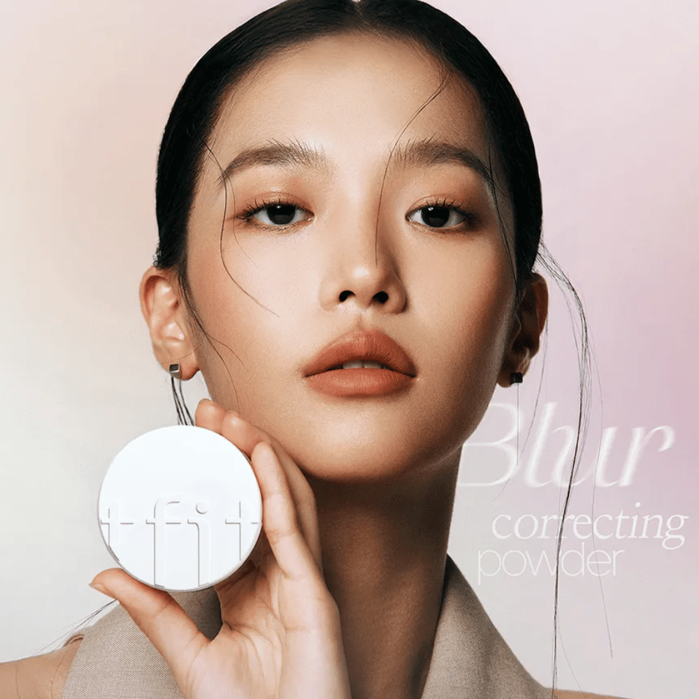 Kvinne med feilfri sminke holder opp en rund hvit boks med TFIT-logo, med teksten "Blur Correcting Powder" i bakgrunnen.