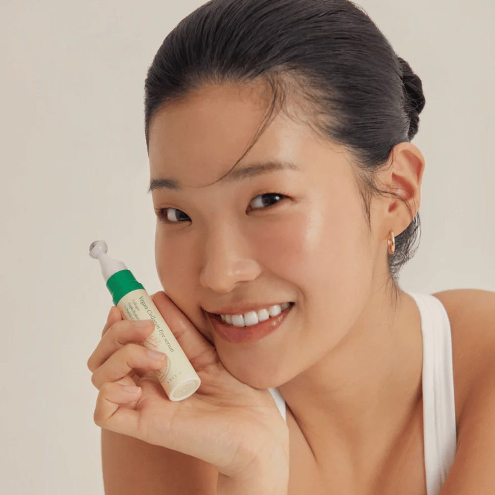 En kvinne med oppsatt hår og gyldne øredobber holder en AXIS-Y Vegan Collagen Eye Serum 10ml nær ansiktet, mens hun smiler mykt. Serumet er designet for å fukte og redusere rynker rundt øynene, spesielt utviklet for sensitiv hud.