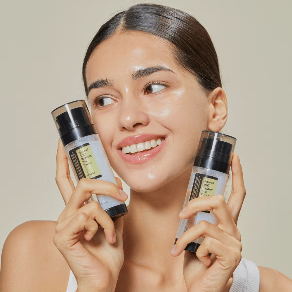 Smilende kvinne med glansfull hud holder to flasker COSRX Advanced Snail Radiance Dual Essence 80 ml nær ansiktet mot en lys bakgrunn.