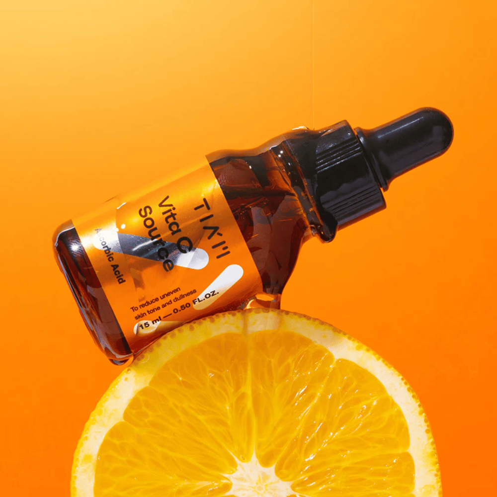 TIA'M Vita C Source-flaske plassert mot en appelsin og en oransje bakgrunn som symboliserer C-vitaminets friskhet og antioksidative effekt.