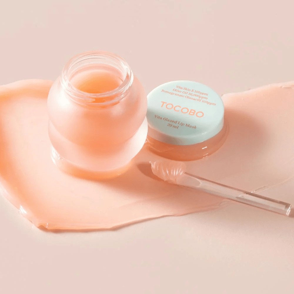 En krukke med "Vita Glazed Lip Mask" fra Tocobo vises. Krukken er gjennomsiktig med et lyst turkist lokk og inneholder en glansfull, rosa leppemaske. En liten spatel ligger ved siden av krukken, og en del av masken er spredt utover underlaget. Produktet er designet for å fukte og pleie leppene.