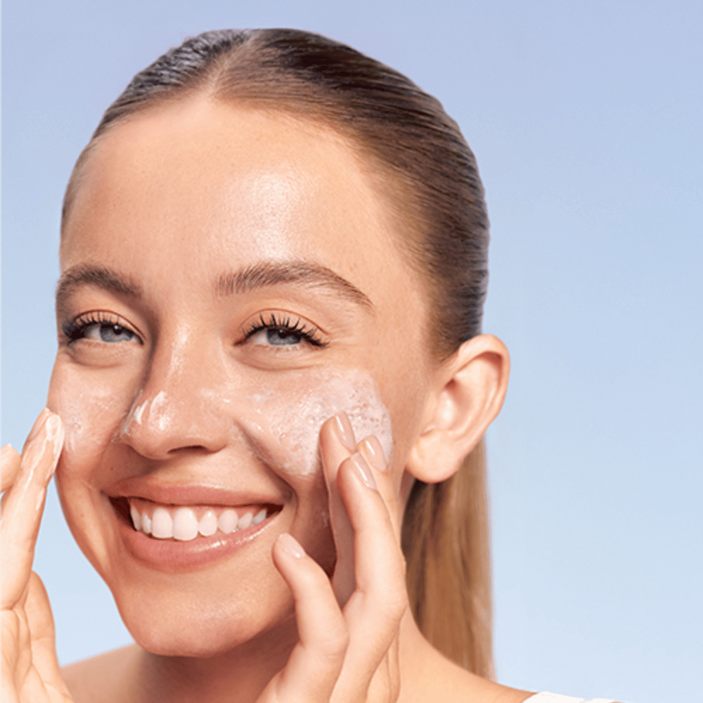Smilende kvinne påfører LANEIGE Water Bank Gentle Gel Cleanser i skummende form på kinnene, mot blå bakgrunn.