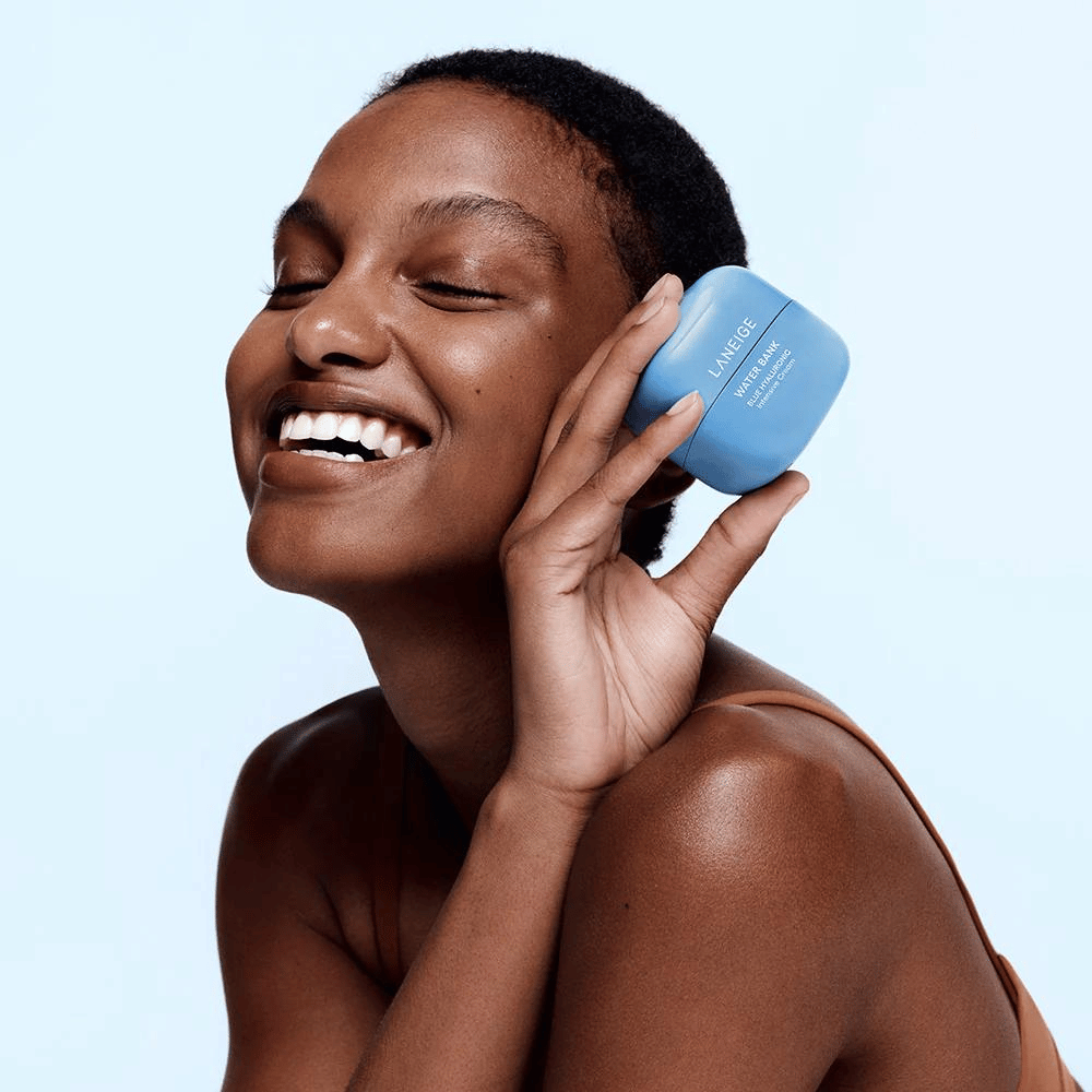 Smilende kvinne holder opp en blå boks LANEIGE Water Bank Blue Hyaluronic Cream mot kinnet, med lyseblå bakgrunn.