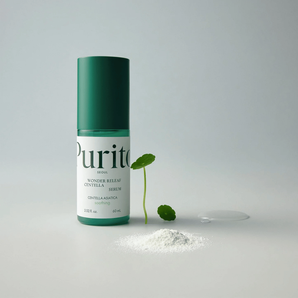 Grønn flaske av Purito Wonder Releaf Centella Serum 60 ml står på et hvitt underlag, omgitt av centellablader, en gjennomsiktig serumsdråpe og et lite hvitt pulver.
