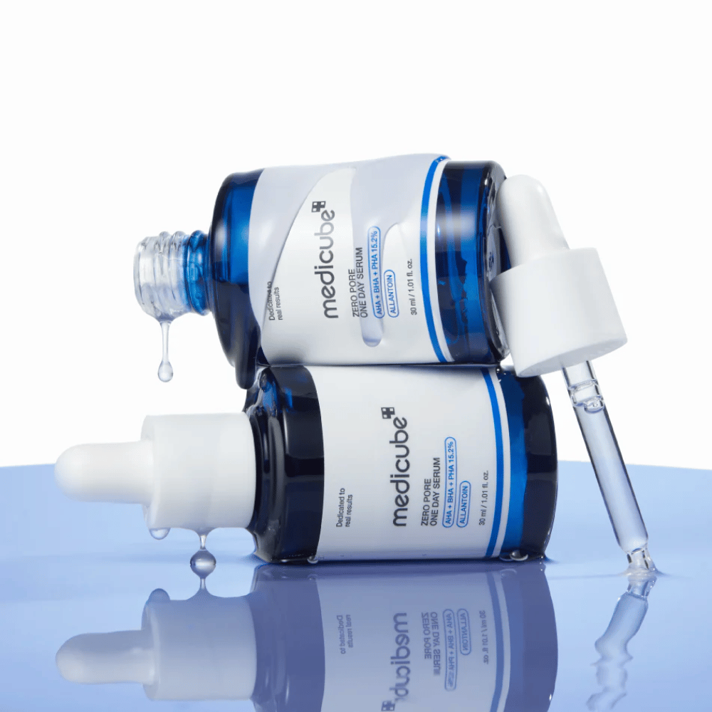 To blå flasker av Medicube Zero Pore One Day Serum liggende mot hverandre, med pipetter og dråper av serum på en reflekterende blå overflate.