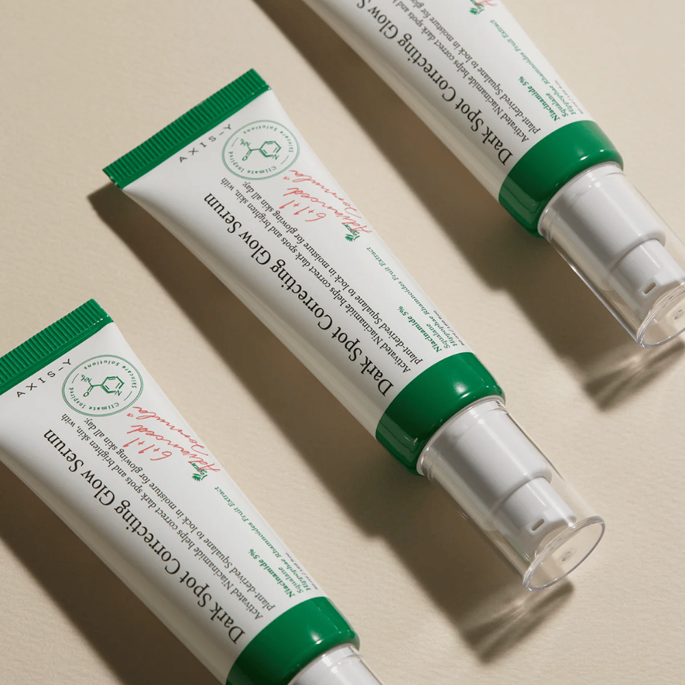 Tre tuber av Axis-Y Dark Spot Correcting Glow Serum med grønn kork og hvit emballasje ligger på et lyst underlag.