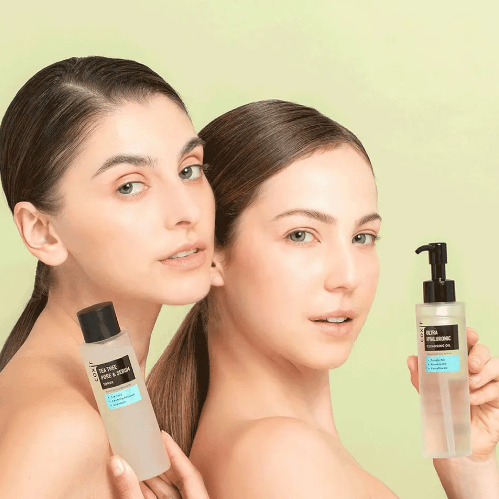 To kvinner med bare skuldre poserer med hudpleieprodukter; den ene holder en flaske merket "TEA TREE PORE & SEBUM", og den andre en pumpeflaske merket "ULTRA HYALURONIC CLEANSING OIL". De står foran en lysegrønn bakgrunn.