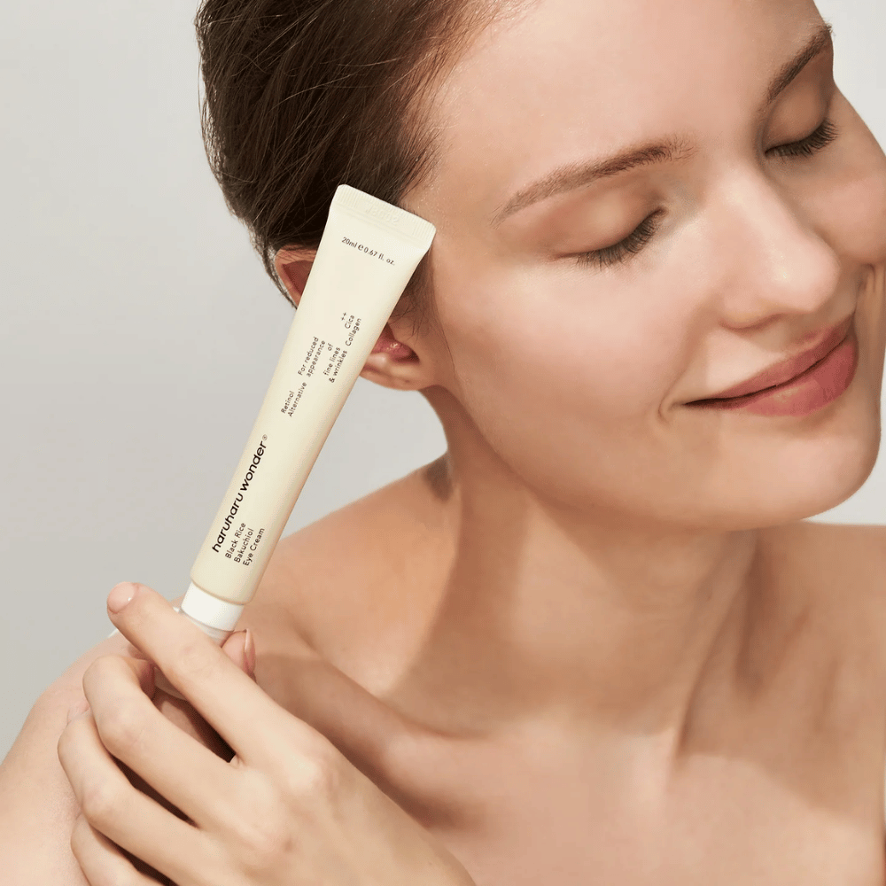 En smilende person med lukkede øyne holder opp en tube Haruharu Wonder Black Rice Bakuchiol Eye Cream mot tinningen.
