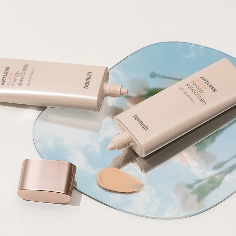 En flaske heimish Artless Glow Tinted Sunscreen SPF50+ PA+++ 40ml og en flaske concealer på et speil, som viser ulike hudtoner.