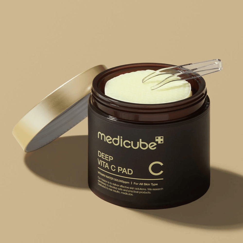 En boks med medicube Deep Vita C Pad. Boksen er mørkebrun med gulltekst og en hvit, invertert etikett med C-vitamin informasjon. Lokket er gull på innsiden, og en gjennomsiktig pinsett ligger oppå de lysegule bomullspadene inni. Bakgrunnen er ensfarget beige.