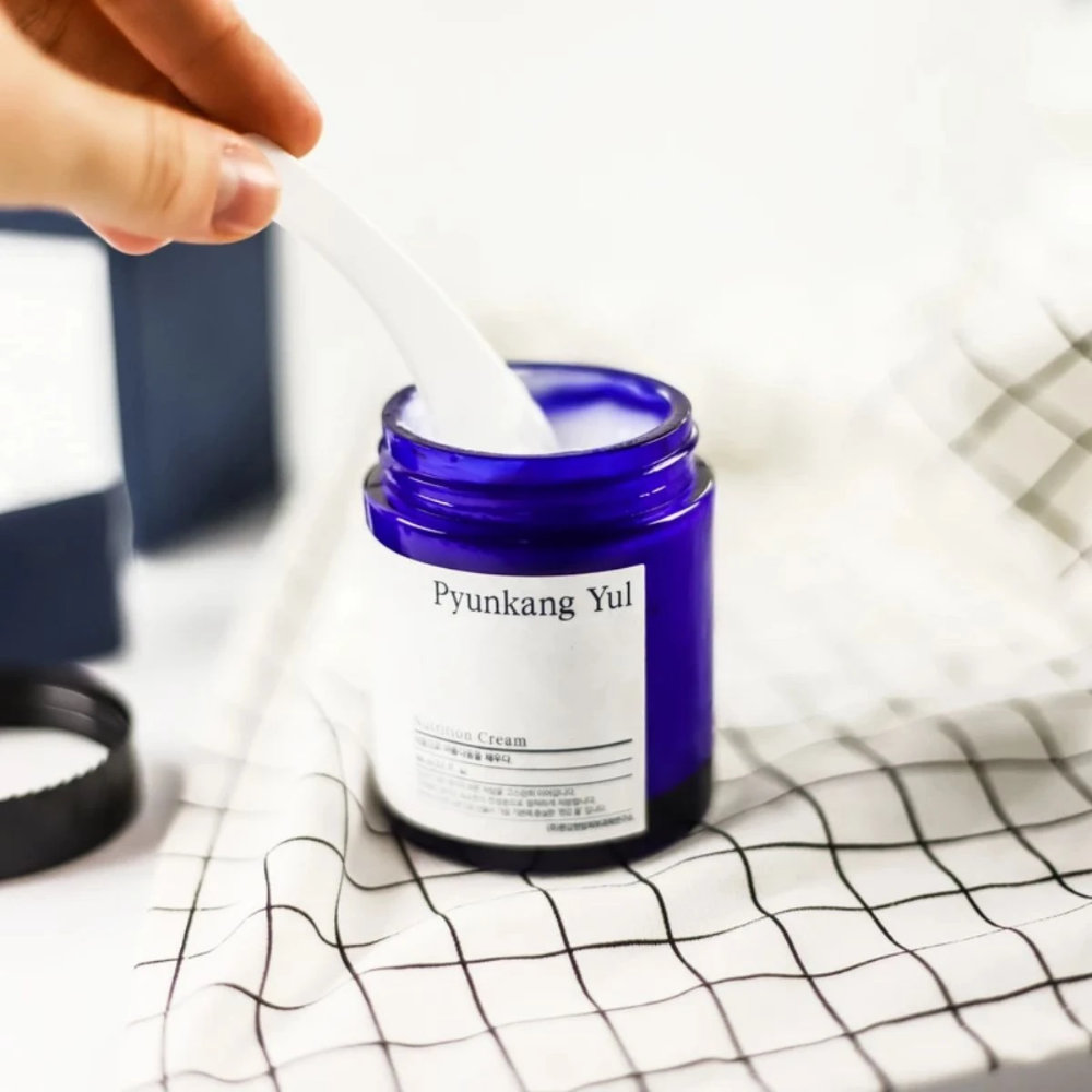 En person bruker en boks Pyunkang Yul Nutrition Cream 100ml på et bord.