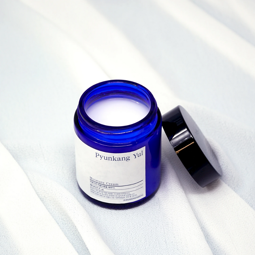 En blå boks med lokk som sitter på en hvit klut som viser frem Pyunkang Yul's Moisture Cream 100ml fuktighetsgivende egenskaper.