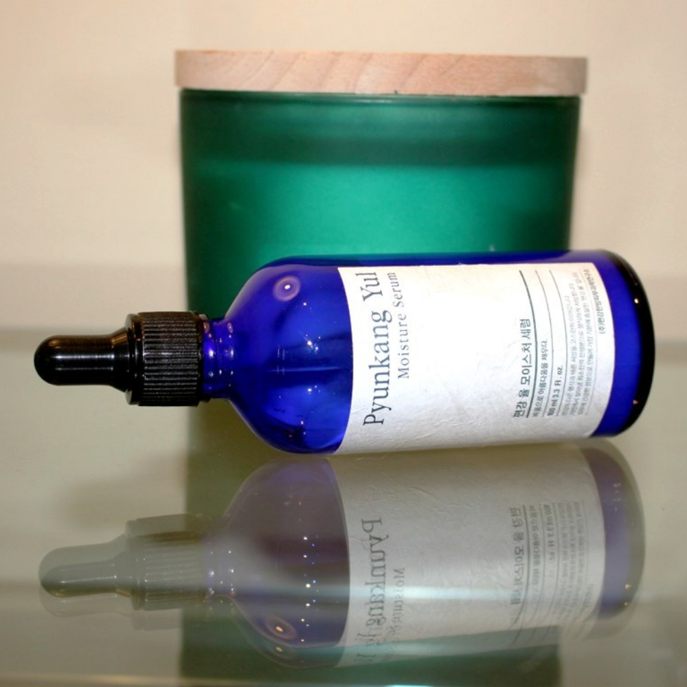 En flaske Pyunkang Yul Moisture Serum 100ml balanserer fuktighetsnivåene på et bord ved siden av et lys.