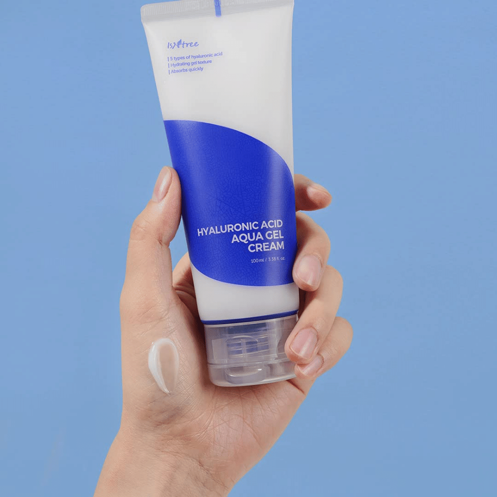 En hånd som holder en tube Isntree Hyaluronic Acid Aqua Gel Cream 100ml på en blå bakgrunn.